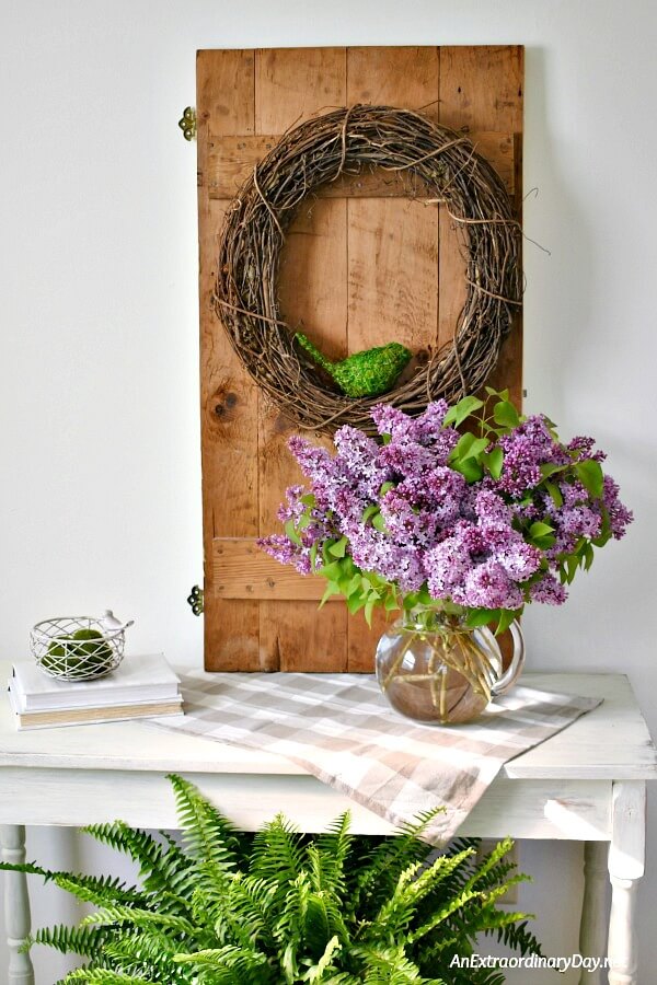 Create pretty seasonal home decor for spring with beautiful vignette showcasing an old door, a grapevine wreath, and a huge bouquet of lilacs. It makes a lovely rustic farmhouse style decor statement. - AnExtraordinaryDay.net