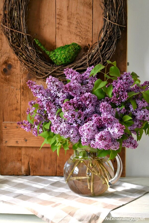 Create pretty seasonal home decor for spring with beautiful vignette showcasing an old door, a grapevine wreath, and a huge bouquet of lilacs. It makes a lovely rustic farmhouse style decor statement.