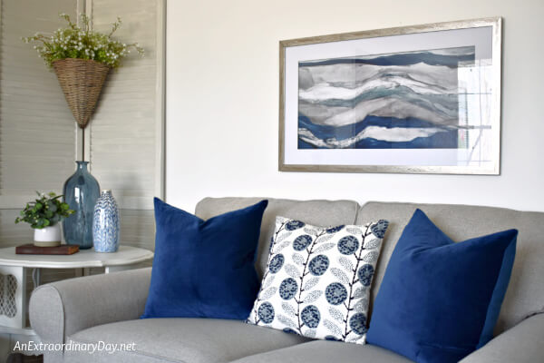 Gray loveseat with dark blue pillows and coordinating painting on the wall of this small apartment living room - AnExtraordinaryDay.net